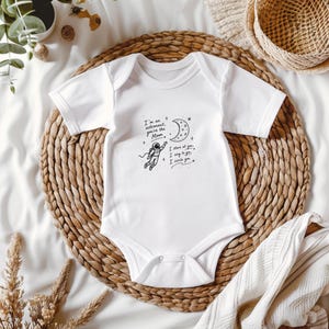 May include: A white short-sleeved baby bodysuit featuring a black line art graphic of an astronaut reaching toward a crescent moon. The text reads: I'm an astronaut, you're the Moon. I stare at you, I sing to you, I circle you. The garment has a crew neckline.