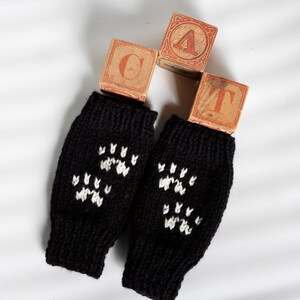 May include: A pair of black knitted fingerless gloves with white paw print designs. The gloves are displayed with three wooden blocks spelling out the word "CAT" in orange letters. The background is a plain white surface.