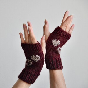 May include: A pair of burgundy fingerless gloves with a knitted heart design in a lighter color. The gloves are made of a soft, textured yarn and are designed to keep hands warm while leaving fingers free.