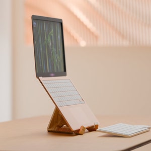 May include: A rose gold laptop is displayed on a wooden stand, showing a bamboo forest image on the screen. A matching wireless keyboard is placed nearby. The setup is on a light brown table.