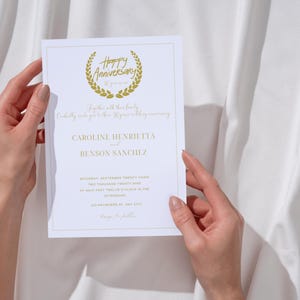 May include: A white wedding invitation with gold lettering. The invitation reads "Happy Anniversary" with a gold laurel wreath. The names "Caroline Henrietta and Benson Sanchez" are listed below the wreath. The date and time of the wedding are listed below the names.