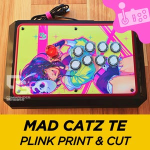 May include: A Mad Catz TE arcade stick with a custom printed faceplate featuring a colourful anime-style illustration of a woman with pink and purple hair. The faceplate has a yellow background with pink and white accents. The arcade stick has eight buttons and a joystick.
