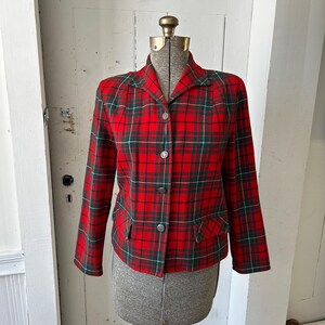 May include: A red plaid blazer with green and white accents. The jacket has a collared neckline, three silver buttons, and two front pockets. The blazer is displayed on a mannequin.