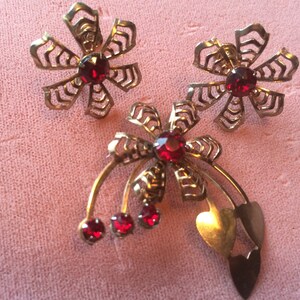 May include: A set of three gold-tone jewelry pieces featuring red rhinestone accents. The set includes two flower-shaped earrings and a brooch with a heart-shaped pendant.