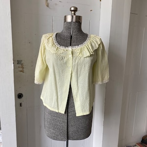 May include: A pale yellow, short-sleeved button-up top with a ruffled lace collar. The top is made of a lightweight, textured fabric.