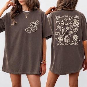 May include: A charcoal gray short-sleeve t-shirt featuring a white line-art locket graphic on the front with the text Mrs. Robert. The back displays wedding-themed illustrations including a chapel, cake, and the text going to the Chapel gonna get married.