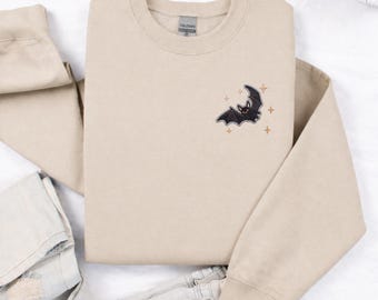 May include: A beige crewneck sweatshirt featuring an embroidered design on the upper left chest. The embroidery depicts a small black bat with outstretched wings surrounded by several thin, orange-colored four-pointed star shapes.