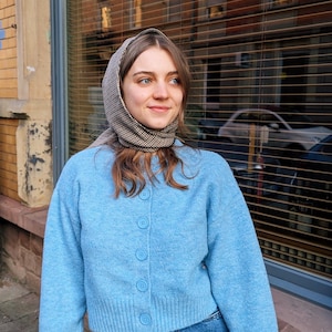 May include: A woman wearing a light blue cardigan with a checkered scarf and blue jeans. The cardigan has a button closure and the jeans are high-waisted.
