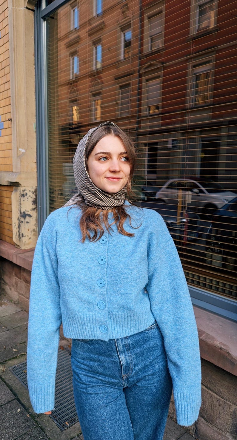 May include: A woman wearing a light blue cardigan with a checkered scarf and blue jeans. The cardigan has a button closure and the jeans are high-waisted.