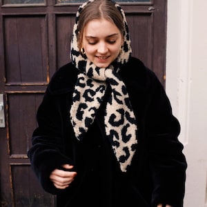 May include: A woman wearing a black coat with a leopard print scarf. The scarf is tied around her neck and the ends are hanging down.