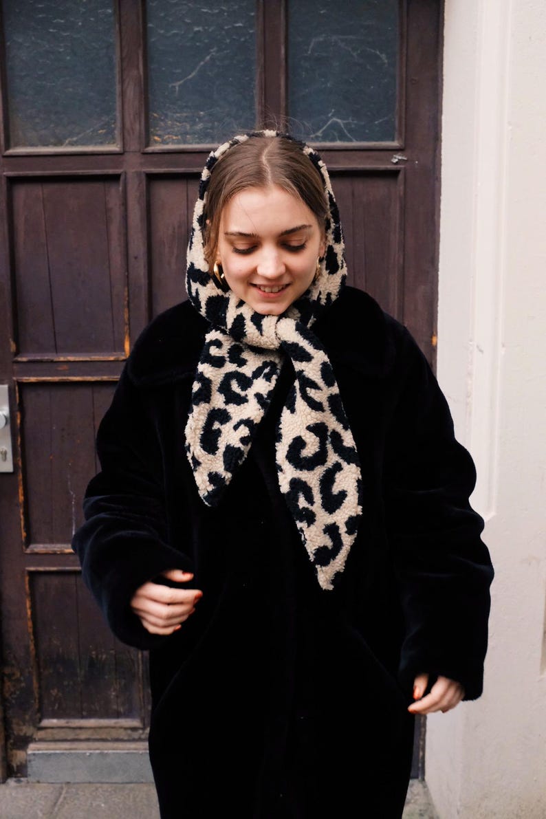 May include: A woman wearing a black coat with a leopard print scarf. The scarf is tied around her neck and the ends are hanging down.