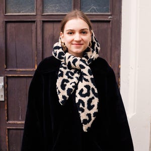 May include: A woman wearing a black coat with a leopard print scarf tied around her neck. She is standing in front of a wooden door.