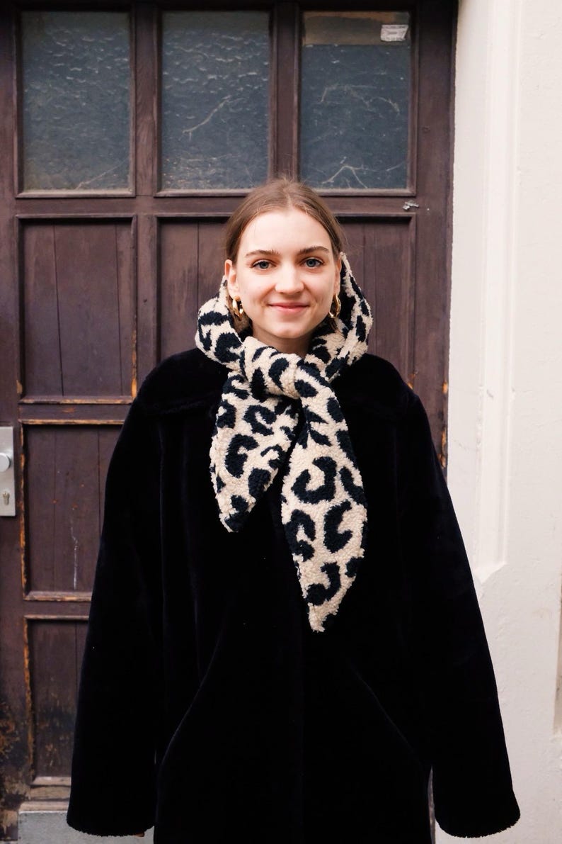 May include: A woman wearing a black coat with a leopard print scarf tied around her neck. She is standing in front of a wooden door.