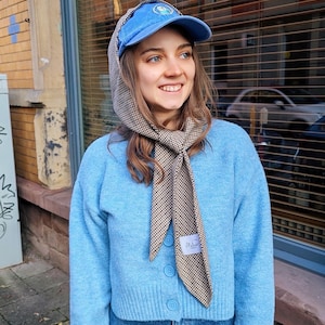 May include: A woman wearing a light blue cardigan with a brown and white checkered scarf tied around her neck. She is wearing a blue baseball cap with a white logo and blue jeans.