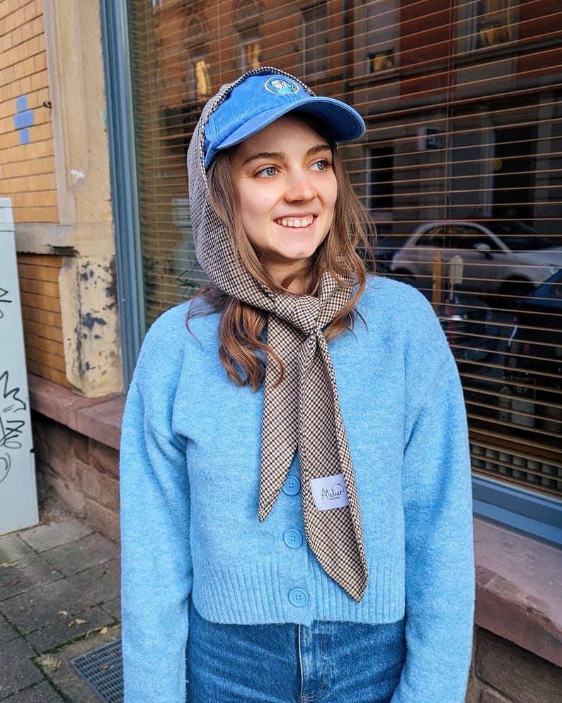 May include: A woman wearing a light blue cardigan with a brown and white checkered scarf tied around her neck. She is wearing a blue baseball cap with a white logo and blue jeans.
