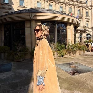 May include: A woman wearing a tan corduroy coat with a leopard print headscarf and sunglasses. She is standing in front of a building with a glass roof.