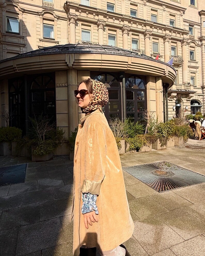 May include: A woman wearing a tan corduroy coat with a leopard print headscarf and sunglasses. She is standing in front of a building with a glass roof.