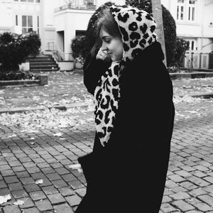 May include: A black and white leopard print hooded scarf with a black coat. The person is walking on a brick pathway.