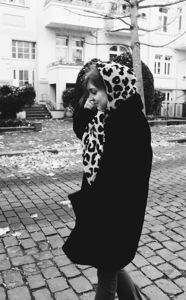 May include: A black and white leopard print hooded scarf with a black coat. The person is walking on a brick pathway.