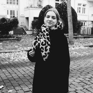 May include: A woman wearing a black coat with a leopard print scarf. She is standing in front of a building with a brick walkway.