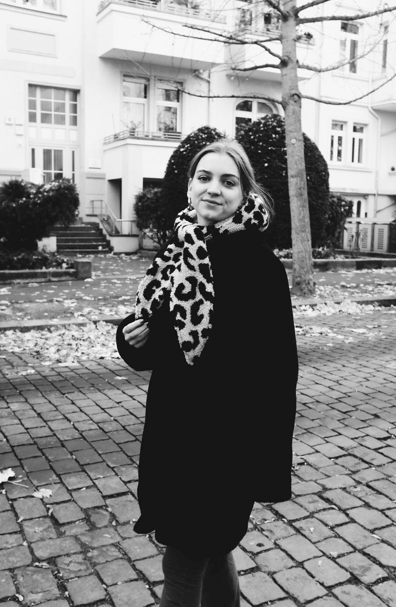 May include: A woman wearing a black coat with a leopard print scarf. She is standing in front of a building with a brick walkway.