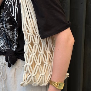 Peut inclure: Un sac cabas en crochet blanc avec un motif noué. Le sac est tenu par une personne portant un t-shirt noir avec un imprimé graphique.
