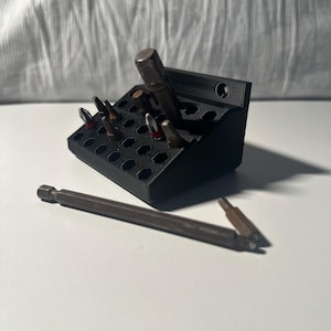 May include: A black plastic organizer with a hexagonal grid for holding screwdriver bits. The organizer has a small hole on the side for mounting. Several screwdriver bits are in the organizer, and two more are lying on a white surface.