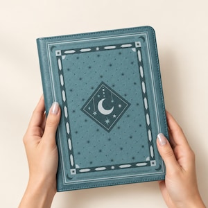 May include: A teal-colored hardcover journal featuring a central diamond-shaped emblem with a crescent moon and star motif. The cover is decorated with a repeating pattern of small stars and a geometric border containing stylized moon and crystal shapes.