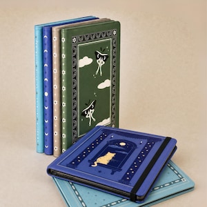May include: A collection of rectangular tablet cases in various colors including light blue, dark blue, beige, and green. The green case features moth and cloud illustrations. A dark blue case in the foreground displays a cat looking out a window.
