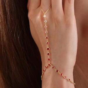 May include: A gold chain bracelet with a finger ring attachment. The bracelet is adorned with small red, pink, and white gemstones.