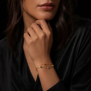 Puede incluir: Pulsera dorada con tres cadenas finas. Cada cadena tiene pequeños amuletos de ojo turco de colores, con detalles en rojo, azul, amarillo y negro. La pulsera se luce en una muñeca sobre un fondo oscuro.