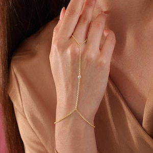 Op de afbeelding: Een gouden kettingarmband met een enkele heldere steentjesaccent. De ketting is verbonden met een ring die aan de vinger kan worden gedragen.