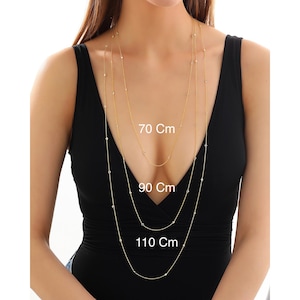 Puede incluir: Tres cadenas de oro con pequeñas piedras transparentes, superpuestas en el cuello de una modelo. Las cadenas están etiquetadas con sus longitudes: 70 cm, 90 cm y 110 cm.