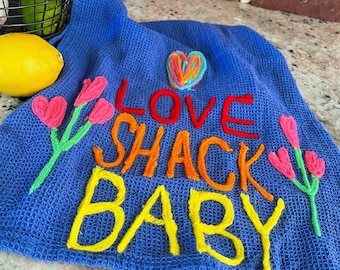 May include: Blue kitchen towel with the text "LOVE SHACK BABY" embroidered in colorful yarn. The towel features a heart and flowers embroidered in pink, green, and yellow yarn.