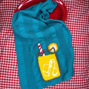 May include: A turquoise kitchen towel with a yellow embroidered cocktail design, including a straw and lemon slice. The towel is folded inside a red bowl, set against a red and white checkered background.
