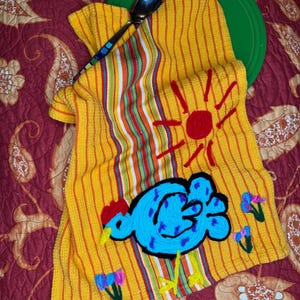 May include: A yellow and orange striped kitchen towel with a blue chicken design, red sun, and colorful flowers. A green plate and a spoon with a patterned handle are also visible.