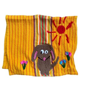 May include: Yellow, textured, rectangular towel with a brown dog, sun, and flowers. The dog has pink ears and a pink nose. The sun is red, and the flowers are pink and blue. The towel has vertical stripes.