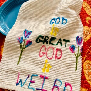 May include: White, textured towel with embroidered text reading "GOD IS GREAT GOD IS WEIRD" in blue, green, pink, and yellow. Features embroidered flowers in pink and blue. Resting on a blue plate.