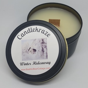 May include: A black tin candle with a lid featuring the text "Candlekraze" and "Winter Hideaway." The candle has a wooden wick and a creamy wax. The lid has a picture of a snowy cabin.