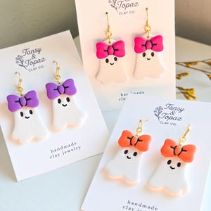 May include: Handmade clay ghost earrings with gold-colored hooks. Each ghost has a smiling face and a colorful bow in purple, pink, or orange. The earrings are displayed on white cards with the text "Tansy & Topaz CLAY CO." and "handmade clay jewelry."