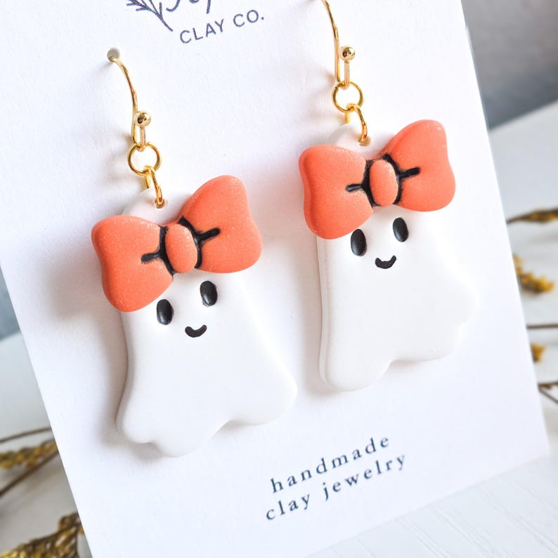 May include: A pair of handmade clay earrings featuring white ghost figures with orange bows and black facial details. The earrings hang from gold-colored hooks. The card behind the earrings reads "handmade clay jewelry."