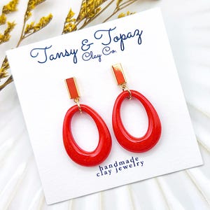 Puede incluir: Pendientes rojos de forma ovalada con partes superiores rectangulares doradas. Se muestran en una tarjeta blanca con el texto "Tansy & Topaz Clay Co." y "handmade clay jewelry". Flores amarillas en el fondo.