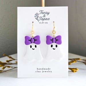 May include: White ghost-shaped earrings with purple bow accents and black facial features. The earrings are handmade clay jewelry with gold-colored ear wires. The card behind the earrings has the text "Tansy & Topaz Clay Co."