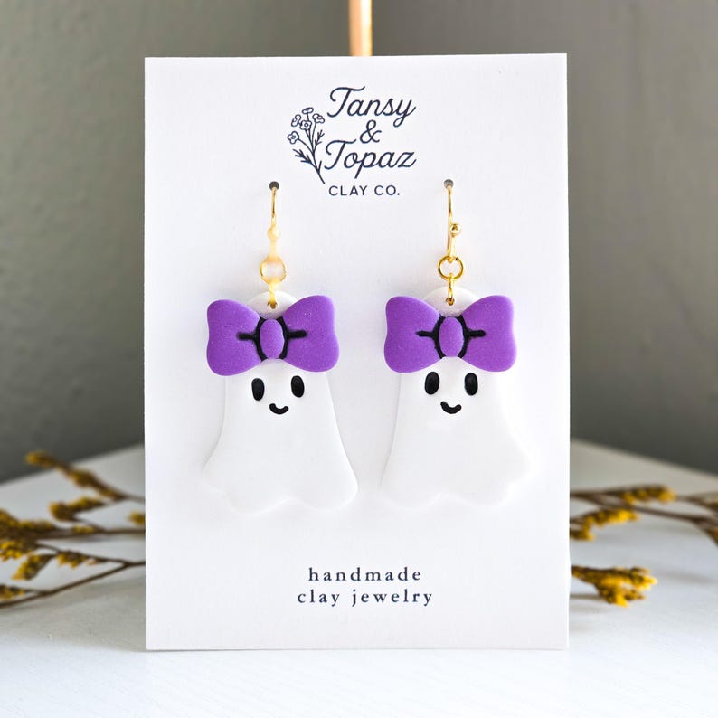 May include: White ghost-shaped earrings with purple bow accents and black facial features. The earrings are handmade clay jewelry with gold-colored ear wires. The card behind the earrings has the text "Tansy & Topaz Clay Co."