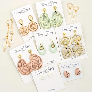 May include: A collection of handmade clay earrings in various designs, including seashell, floral, and geometric shapes. The earrings are in pastel colors like pink, green, and white, with gold accents. Each pair is displayed on a white card with the brand name.