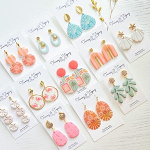May include: A collection of handmade clay earrings in various designs, including leaf, seashell, and geometric patterns. The earrings feature gold-colored hardware and are displayed on white cards with the text "Tansy & Topaz handmade clay jewelry."