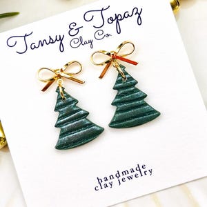 May include: A pair of handmade clay jewelry earrings. Each earring features a green Christmas tree charm with gold-colored bow accents. The earrings are displayed on a white card with the text "Tansy & Topaz Clay Co." and "handmade clay jewelry".