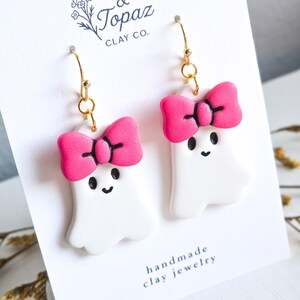 May include: A pair of handmade clay ghost earrings. Each white ghost has a bright pink bow and black facial features. The earrings hang from gold-colored hooks. The card reads "& Topaz Clay Co." and "handmade clay jewelry."