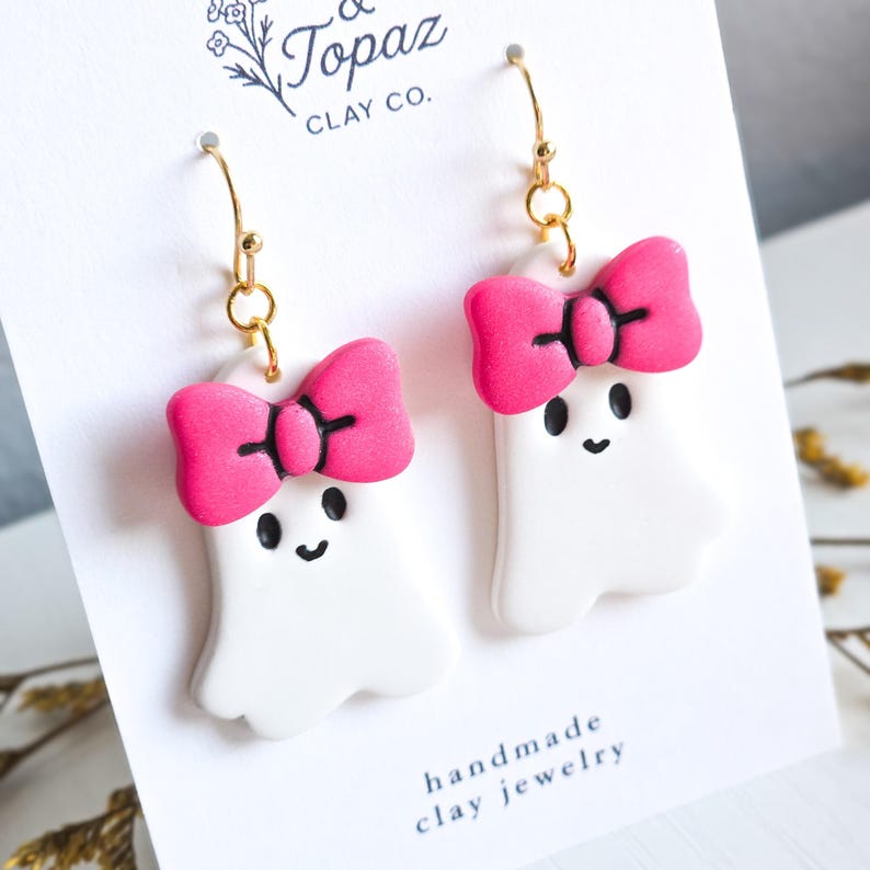 May include: A pair of handmade clay ghost earrings. Each white ghost has a bright pink bow and black facial features. The earrings hang from gold-colored hooks. The card reads "& Topaz Clay Co." and "handmade clay jewelry."