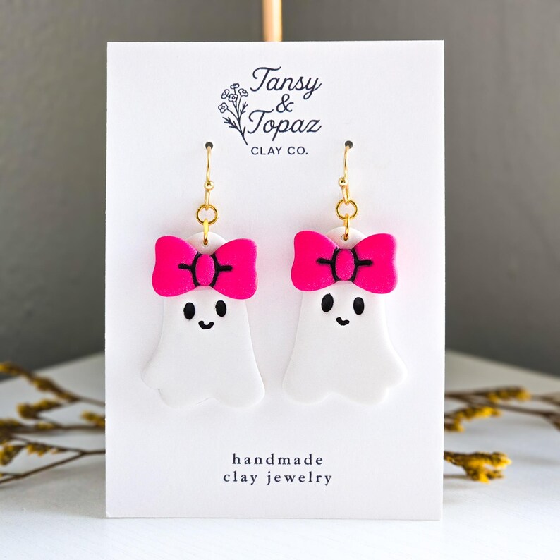May include: White ghost-shaped earrings with bright pink bows and black details. The earrings are handmade clay jewelry with gold-colored hooks. The card behind the earrings reads "Tansy & Topaz Clay Co."